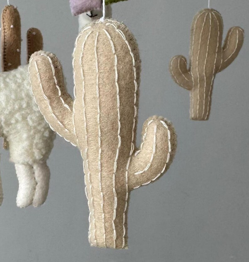 Puede incluir: M&oacute;vil de cactus de fieltro con una paleta de colores neutros. El m&oacute;vil presenta varios cactus de fieltro de diferentes tama&ntilde;os y formas, con detalles de costura blancos. Tambi&eacute;n se ve una llama y otras formas de fieltro.