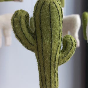 Puede incluir: Un cactus de fieltro verde con l&iacute;neas verticales verde oscuro. El cactus, con dos brazos, forma parte de un m&oacute;vil. Otras formas de fieltro, incluido un llama blanco, son visibles en el fondo. Es un elemento decorativo.
