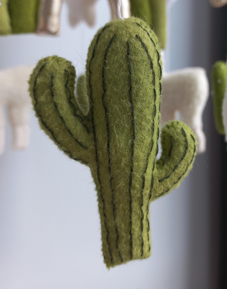 Puede incluir: Un cactus de fieltro verde con l&iacute;neas verticales verde oscuro. El cactus, con dos brazos, forma parte de un m&oacute;vil. Otras formas de fieltro, incluido un llama blanco, son visibles en el fondo. Es un elemento decorativo.