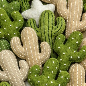 Puede incluir: Un surtido de decoraciones de cactus de fieltro en varios tonos de verde y beige. Los cactus tienen detalles de costura blancos y peque&ntilde;os puntos blancos. Tambi&eacute;n hay una decoraci&oacute;n en forma de estrella blanca, creando un arreglo l&uacute;dico con tem&aacute;tica des&eacute;rtica.