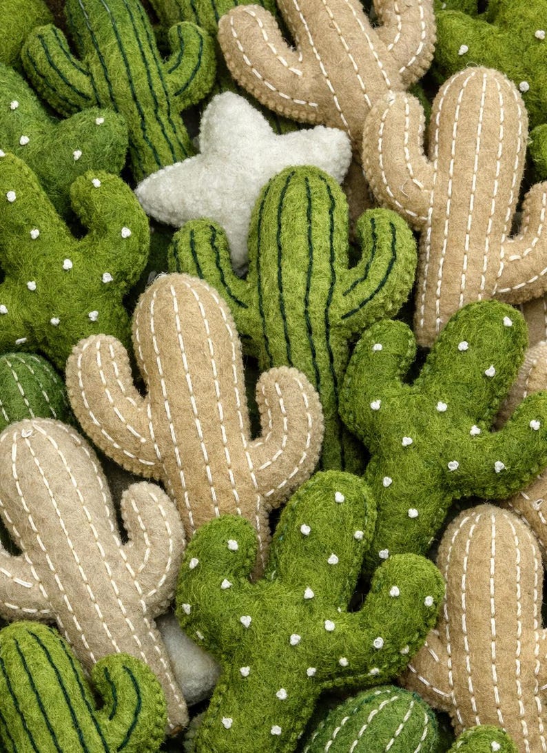 Puede incluir: Un surtido de decoraciones de cactus de fieltro en varios tonos de verde y beige. Los cactus tienen detalles de costura blancos y peque&ntilde;os puntos blancos. Tambi&eacute;n hay una decoraci&oacute;n en forma de estrella blanca, creando un arreglo l&uacute;dico con tem&aacute;tica des&eacute;rtica.