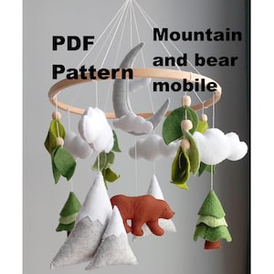Puede incluir: Móvil para bebé hecho a mano con montañas, árboles, nubes, un oso y una luna creciente de fieltro. El móvil está suspendido de un aro de madera y contiene el texto "PDF Pattern Mountain and bear mobile".