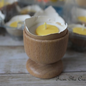 May include: A wooden egg cup contains a candle designed to resemble a fried egg. The candle features a yellow yolk and a white 'egg white' edge. Other similar candles are visible in the background. The text "From The Craft Room" is at the bottom.