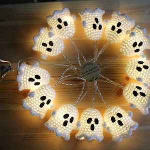 May include: A string of ten white crocheted ghost-shaped light covers arranged in a circle. Each ghost features black felt eyes and a mouth. The lights are connected by a thin wire to a clear plastic battery pack containing two AA batteries.