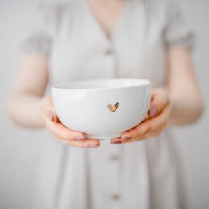 Op de afbeelding: Een witte keramische kom met een gouden hartontwerp. De kom wordt vastgehouden door de handen van een persoon.