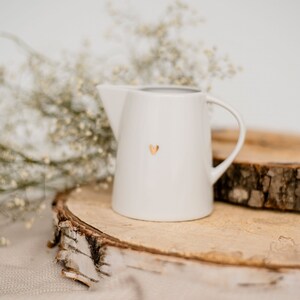 Peut inclure: Pichet en céramique blanche avec un motif de cœur doré. Le pichet est posé sur une tranche de bois.