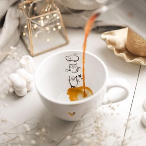 Op de afbeelding: Een witte keramische mok met een zwarte lijntekening van drie katten. De mok is gevuld met koffie en de koffie wordt in de mok gegoten.