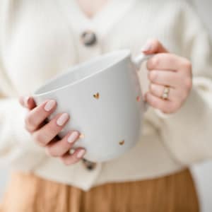 Könnte beinhalten: Eine weiße Keramiktasse mit goldenen Herzmotiven. Die Tasse wird von den Händen einer Person gehalten.