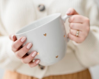 May include: A white ceramic mug with gold heart designs. The mug is being held by a person's hands.