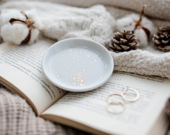 Op de afbeelding: Een witte keramische ringenschotel met gouden vlekken op een wit boek met tekst in het Pools. De schotel ligt op een wit gebreid deken met wattenbolletjes en dennenappels.