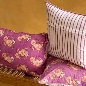 May include: Three decorative throw pillows with floral patterns. The pillows are pink and white with a floral print. The pillows are arranged on a wooden surface.