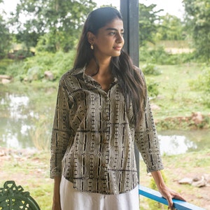 Puede incluir: Una mujer con una camisa de estampado blanco y negro con cuello blanco y mangas largas. Está de pie frente a una ventana con vista a un estanque y vegetación.