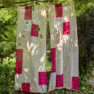 Könnte beinhalten: Zwei transparente, cremefarbene Vorhänge mit einem Patchwork-Design. Das Patchwork umfasst weißen Stoff mit kleinen Punkten und einfarbigen, pinkfarbenen Rechtecken. Die Vorhänge hängen an einem Ast, mit Grün im Hintergrund.