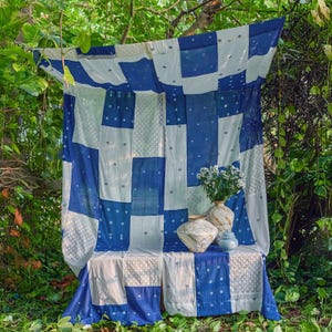 May include: A blue and white patchwork textile is draped over a bench, creating a backdrop. The textile features large squares of solid blue and white patterned fabric. Vases with flowers sit on the bench. The scene is set outdoors, surrounded by greenery.