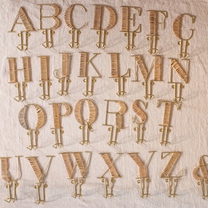 May include: A set of 26 gold-colored metal letters, each shaped like a letter of the alphabet, with a hook at the bottom. The letters are arranged in alphabetical order, and the background is a light beige fabric.