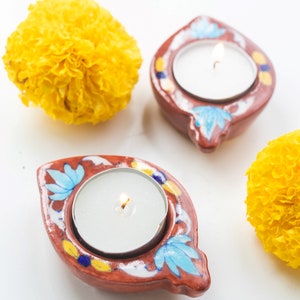 May include: Two terracotta candle holders with blue and yellow floral designs and lit tea lights. The holders are on a white surface with two yellow marigold flowers.