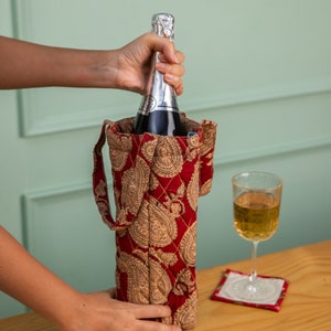 May include: A red and gold paisley patterned fabric wine bottle carrier with a bottle of champagne being inserted into the carrier.