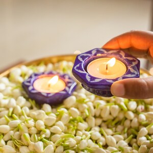 May include: Two blue and white ceramic candle holders with lit tea lights, surrounded by white jasmine flower buds in a gold bowl.