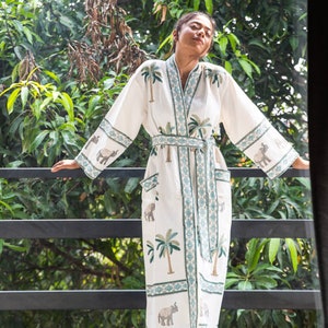 May include: White cotton robe with a green and blue embroidered pattern of palm trees and elephants. The robe has a tie belt and long sleeves.