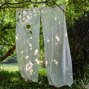 Puede incluir: Dos cortinas blancas transparentes cuelgan de una rama de árbol, iluminadas por la luz del sol que se filtra a través de las hojas. Las cortinas presentan un diseño de patchwork con varios patrones geométricos, incluyendo chevrones y lunares, creando una estética delicada y aireada.