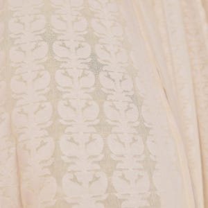 K&ouml;nnte beinhalten: Nahaufnahme eines cremefarbenen Stoffes mit einem sich wiederholenden Blumenmuster. Das Muster zeigt symmetrische Blumenmotive in einem helleren Cremeton, wodurch ein subtiler Kontrast entsteht. Der Stoff wirkt weich und flie&szlig;end, mit sichtbarer Textur.