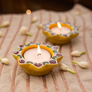 May include: Two yellow ceramic candle holders with floral designs and white tea lights burning inside. The candle holders are on a striped fabric surface.