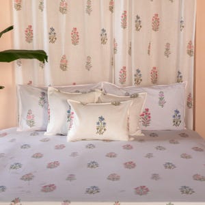 Puede incluir: Una cama blanca con un edredón de color morado claro con estampado floral y fundas de almohada blancas. Las almohadas tienen un diseño floral en tonos rosa, azul y verde. La cama está contra una cortina blanca con un estampado floral.
