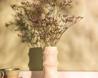 May include: A white ceramic vase with a textured surface, resembling bamboo, holds a bouquet of light purple flowers. The vase is sitting on a woven mat.