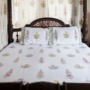May include: White bed linen with a floral print in shades of pink, yellow and green. The bed is made with two pillows and a wooden headboard.