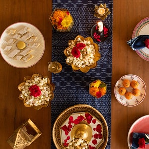 May include: A table setting with a blue and white patterned tablecloth. There are several plates with different types of Indian sweets, flowers, candles, and other decorative items.