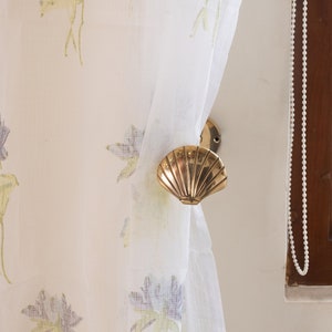 Peut inclure: Un embrasse de rideau en forme de coquille de couleur or. L'embrasse est fixée à un rideau blanc transparent à motif floral.