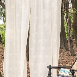 May include: A pair of off-white curtains featuring a subtle, repeating square grid pattern woven into the fabric. The curtains hang in vertical folds, displaying a semi-sheer texture that allows light to pass through the material.