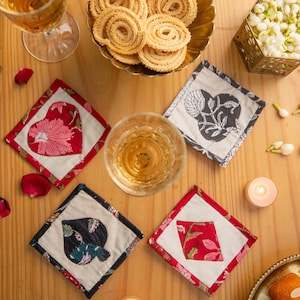 Puede incluir: Cuatro posavasos cuadrados de tela con diferentes patrones. Los posavasos son rojos, blancos y negros con diseños florales. Los posavasos están sobre una superficie de madera.