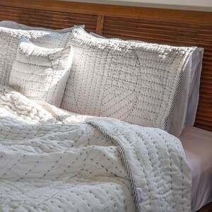 May include: White cotton quilt with black stitching in a geometric pattern. The quilt is on a bed with white pillowcases and a wooden headboard.