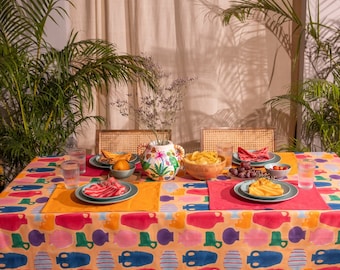 May include: A vibrant table setting with a colorful tablecloth featuring a repeating pattern of abstract shapes in various colors. The table is set with plates, glasses, and a decorative vase filled with flowers. Snacks and food are arranged on the table.