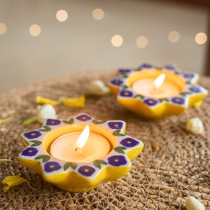May include: Two yellow star-shaped candle holders with purple floral designs and white accents. Each holder has a small white tea light candle inside, with the flames lit.