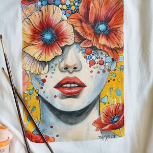 May include: A hand-painted t-shirt featuring a colourful illustration of a woman's face with red poppies covering her eyes. The background is yellow with small colourful dots and the artist's signature "DAXXSArt" is in the bottom right corner.