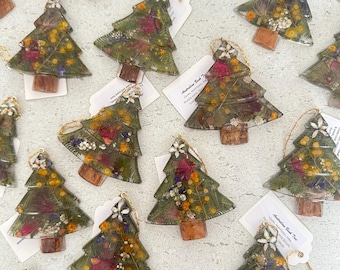 May include: A collection of Christmas tree-shaped ornaments made of clear resin, each containing pressed dried flowers, green foliage, and a small wooden trunk base. Each ornament features a gold hanging string and a white paper tag.