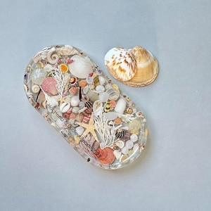 May include: An oval resin tray filled with seashells, starfish, and small stones. The tray has the text "Hot Head & Dudley Beach 2023". Two seashells are next to the tray. The tray and shells are on a light blue background.