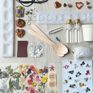 May include: A resin jewellery making kit with dried flowers, moulds, resin, and tools. The kit includes two bottles of resin, a silicone mould with heart shapes, a wooden spoon, a pair of tweezers, a small bag of glitter, a sanding block, a small cup, a bag of dried flowers, and two stickers that say "Hand Made".