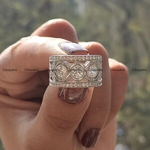 May include: A silver ring with three large round stones surrounded by smaller stones. The ring is set in a wide band with a decorative design.