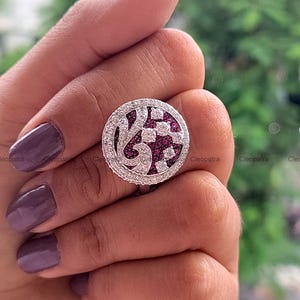 May include: A circular silver ring with a central design of white and red stones. The ring is edged with small, clear stones. The ring is held up by a person's hand.