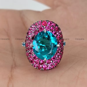 May include: An oval-shaped ring featuring a large, vibrant teal gemstone at its center, surrounded by a halo of smaller pink gemstones. The ring has a silver band and small blue accent stones on each side.