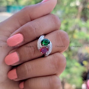 Puede incluir: Un anillo plateado con dos piedras preciosas ovaladas, una verde y otra roja, colocadas una al lado de la otra. El anillo está adornado con pequeñas piedras claras a lo largo de la banda. El anillo es sostenido por una mano con esmalte de uñas rosa.