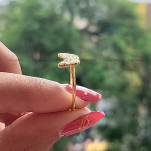 May include: A gold-colored ring featuring a crescent moon design encrusted with small, sparkling stones. The ring is held against a blurred green background, showcasing the jewelry's delicate details and craftsmanship.