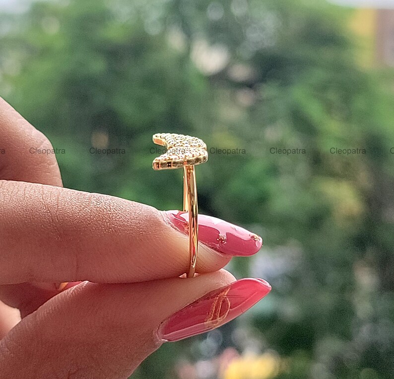 May include: A gold-colored ring featuring a crescent moon design encrusted with small, sparkling stones. The ring is held against a blurred green background, showcasing the jewelry's delicate details and craftsmanship.
