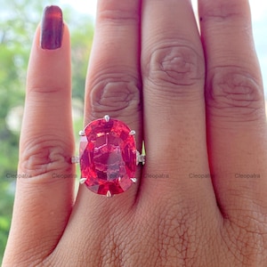 Puede incluir: Un anillo con una gran piedra preciosa de color rojo vibrante y corte ovalado, montada en una banda de metal plateado con cuatro garras. La piedra muestra una superficie facetada que refleja la luz y la palabra Cleopatra aparece repetida en la imagen.