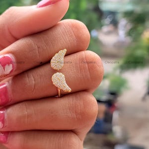 May include: A gold-colored ring with two wing-shaped designs, each encrusted with small, sparkling stones. The ring is open, allowing it to be adjusted to fit different finger sizes. The ring is held by a person.