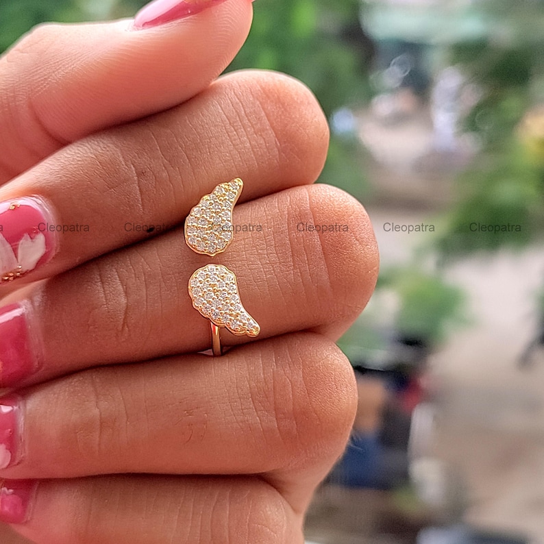 May include: A gold-colored ring with two wing-shaped designs, each encrusted with small, sparkling stones. The ring is open, allowing it to be adjusted to fit different finger sizes. The ring is held by a person.