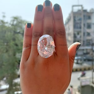 May include: A large oval-cut clear gemstone ring set in a silver-toned metal band. The faceted stone features a prominent brilliant cut that reflects light, secured by small prongs on a simple band worn on the middle finger of a hand.